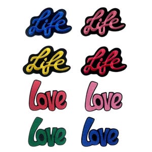 Peut inclure: Plusieurs écussons brodés avec les mots "Life" et "Love" en différentes couleurs. Les écussons "Life" sont bleus, roses, jaunes et rouges. Les écussons "Love" sont rouges, roses, verts et bleus. Chaque écusson a une bordure noire.