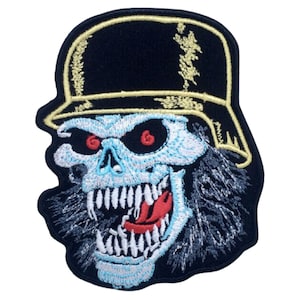 Puede incluir: Parche bordado con una calavera que lleva un casco negro y dorado. La calavera tiene ojos rojos y una expresión amenazante con los dientes descubiertos.
