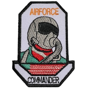 May include: A white embroidered patch with a black outline. The patch features a cartoon image of a pilot wearing a helmet and oxygen mask. The text "AIRFORCE" is in orange above the pilot and "COMMANDER" is below the pilot. The pilot is wearing a green jumpsuit with a red and white scarf.