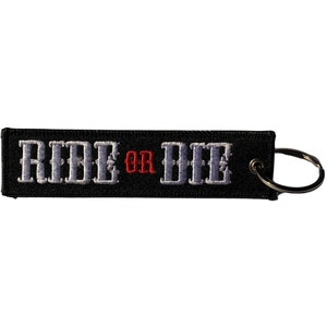 Könnte beinhalten: Schwarzer Schlüsselanhänger mit einem weißen gestickten "RIDE OR DIE"-Text. Das Wort "OR" ist rot. Der Schlüsselanhänger hat einen silbernen Metallring.