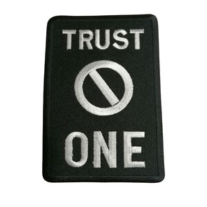 Könnte beinhalten: Ein schwarzer gestickter Aufnäher mit weißen Nähten, auf dem "TRUST NO ONE" mit einem Kreis und einer diagonalen Linie durch ihn hindurch steht.