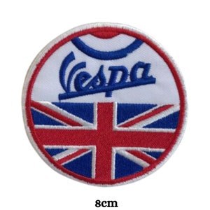 Può includere: Un cerotto rotondo ricamato bianco e blu con la parola "Vespa" in lettere corsive blu e una bandiera Union Jack rossa, bianca e blu. Il cerotto ha un diametro di 8 cm.