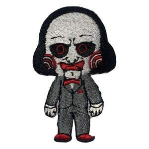 May include: Embroidered iron-on patch featuring a cartoon depiction of the Jigsaw character from the Saw horror film franchise. The character is wearing a black suit and has a red and white spiral pattern on his face.
