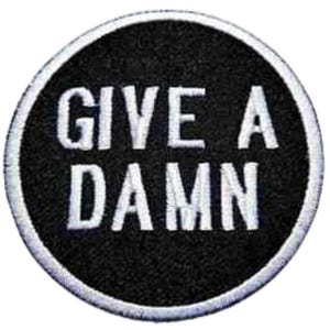 Op de afbeelding: Ronde zwarte patch met witte geborduurde tekst "GIVE A DAMN". De patch heeft een witte rand. Deze patch kan op kleding of accessoires worden genaaid of gestreken.