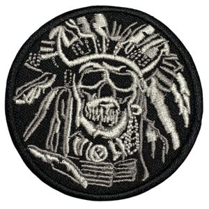 May include: Embroidered patch featuring a skull wearing a Native American headdress. The design is in shades of gray and white against a black circular background. The patch is likely for clothing or accessories.