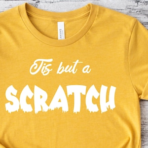 Peut inclure: T-shirt jaune moutarde avec l'inscription blanche « Tis but a Scratch » en texte stylisé. Le t-shirt est noué en bas. Le t-shirt est sur un fond de grain de bois clair.