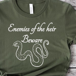 Puede incluir: Una camiseta verde con un gráfico de serpiente blanca y el texto "Enemies of the heir Beware".