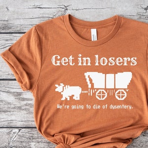 May include: A terracotta-coloured t-shirt featuring the phrase "Get in losers" above a pixelated wagon and ox. The text "We're going to die of dysentery" is below the wagon.