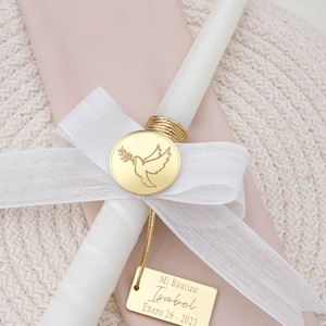 May include: A white baptism candle with a gold dove and olive branch charm. The candle is tied with a white ribbon and has a gold tag that reads "Mi Bautizo Isabel Enero 26, 2023".