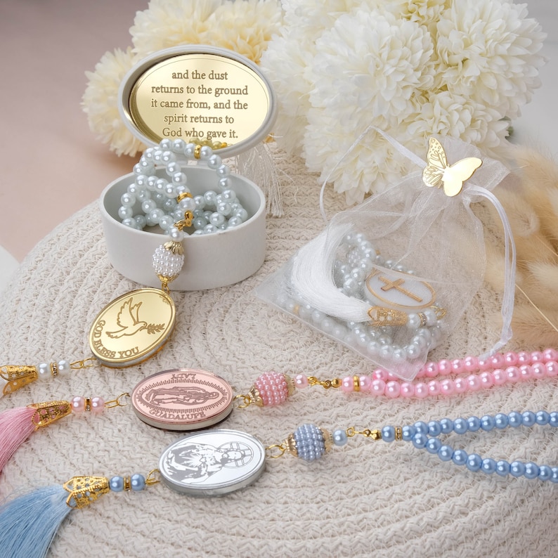 May include: Three sets of prayer beads, each with a different charm. The beads are white, pink, and blue. The charms are a dove, a religious figure, and a baby Jesus. The beads are in a white box with a gold inscription that reads "and the dust returns to the ground it came from, and the spirit returns to God who gave it."