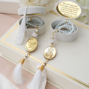May include: Two white boxes with gold trim, each containing a white pearl necklace and a gold pendant. One pendant features a dove with the words "God Bless You" and the other features a religious image. The boxes are on a pink and white surface. The text on the box reads "Be strong and courageous for the Lord your God will be with you wherever you go Joshua 1:9."