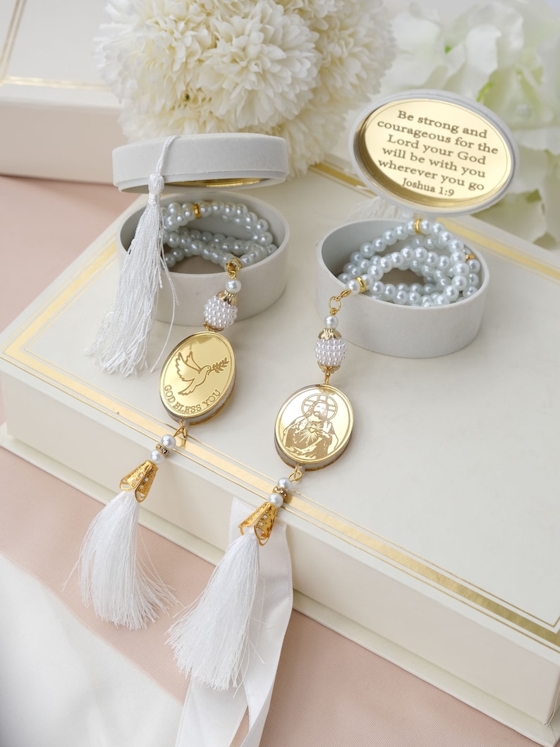 May include: Two white boxes with gold trim, each containing a white pearl necklace and a gold pendant. One pendant features a dove with the words "God Bless You" and the other features a religious image. The boxes are on a pink and white surface. The text on the box reads "Be strong and courageous for the Lord your God will be with you wherever you go Joshua 1:9."