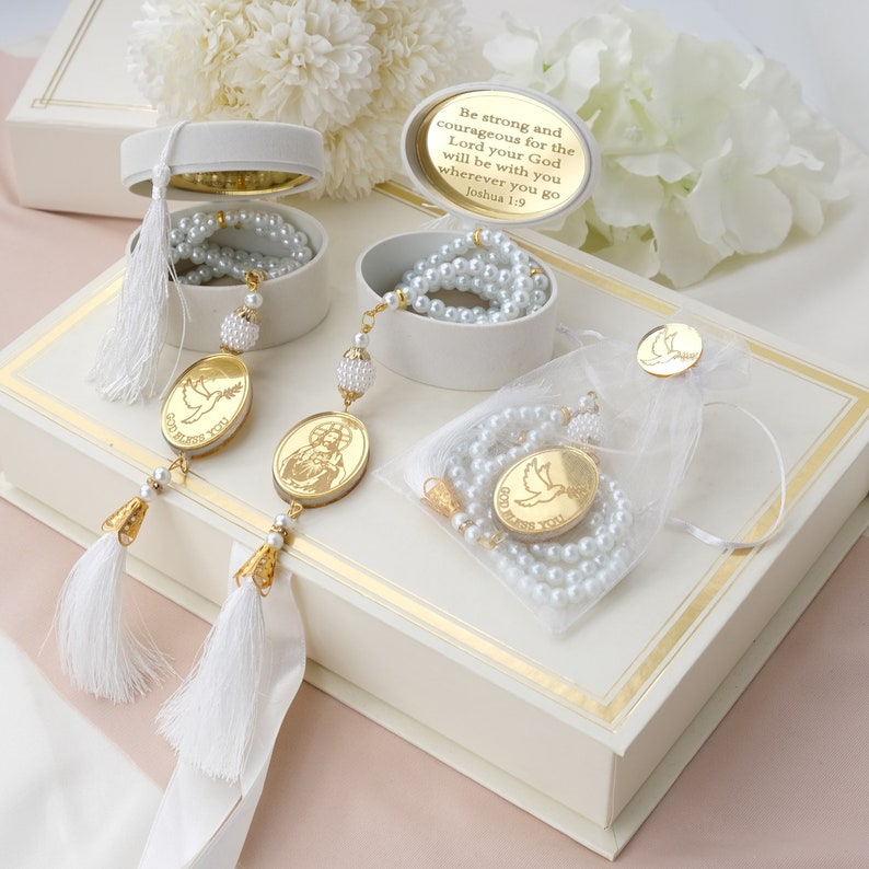 May include: A white box with gold trim contains three sets of white prayer beads with gold accents. Each set includes a small gold pendant with a religious image. The box lid has a quote from Joshua 1:9, "Be strong and courageous for the Lord your God will be with you wherever you go."