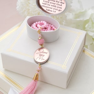 May include: Pink prayer beads with a gold tassel and a round, rose gold charm with the words "Jesus Loves Me" on a white box. The box has a gold trim and a round, white plaque with the text "Be strong and courageous for the Lord your God will be with you wherever you go Joshua 1:9".
