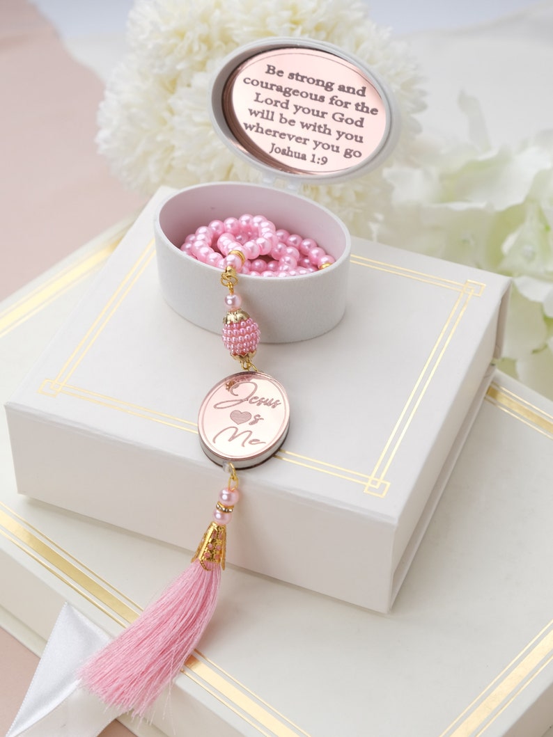 May include: Pink prayer beads with a gold tassel and a round, rose gold charm with the words "Jesus Loves Me" on a white box. The box has a gold trim and a round, white plaque with the text "Be strong and courageous for the Lord your God will be with you wherever you go Joshua 1:9".
