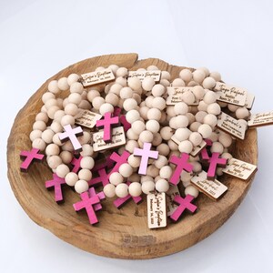 May include: A wooden bowl filled with light-colored wooden bead rosaries and pink crosses. Small wooden tags with names and dates are attached to some of the rosaries. The text on the tags includes names like Sofia and Isabel, and dates like February 26, 2023.