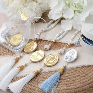 May include: Three white pearl rosaries with gold accents and personalized charms. The charms feature religious imagery and the name "Nikola" with the text "Blessed 6.11.2023". The rosaries are displayed on a burlap surface with white and blue tassels.