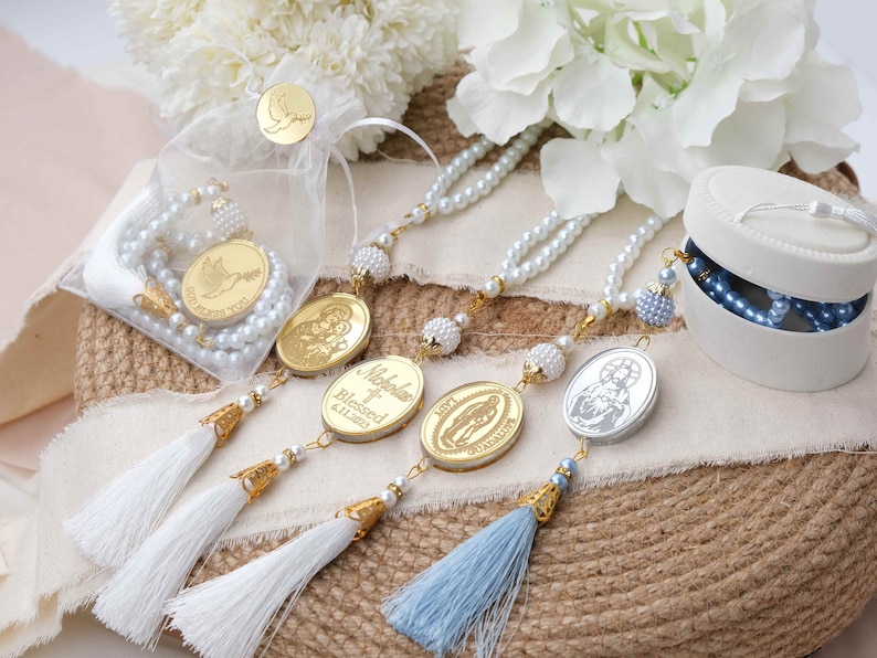 May include: Three white pearl rosaries with gold accents and personalized charms. The charms feature religious imagery and the name "Nikola" with the text "Blessed 6.11.2023". The rosaries are displayed on a burlap surface with white and blue tassels.