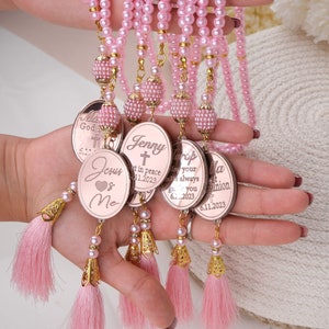 May include: A set of pink pearl rosaries with gold accents and personalized charms. Each charm features a cross, a heart, and a personalized message. The charms are attached to pink tassels.