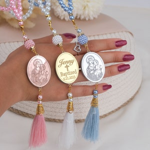 May include: Three gold-toned oval charms with a religious image and a cross, with the name "Jenny" and the date "6.11.2023" engraved on one charm. Each charm is attached to a string of beads with a tassel at the end. The beads are pink, white, and blue.