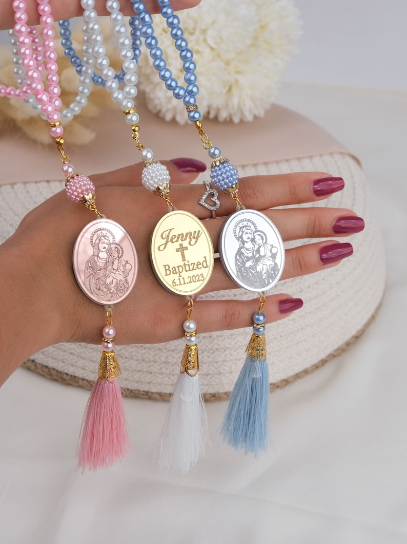 May include: Three gold-toned oval charms with a religious image and a cross, with the name "Jenny" and the date "6.11.2023" engraved on one charm. Each charm is attached to a string of beads with a tassel at the end. The beads are pink, white, and blue.