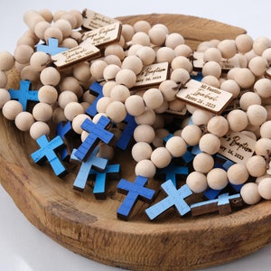 May include: Wooden bead rosaries with blue crosses and personalized tags. The tags are engraved with names and dates, likely for a baptism or first communion. The rosaries are displayed in a wooden bowl.