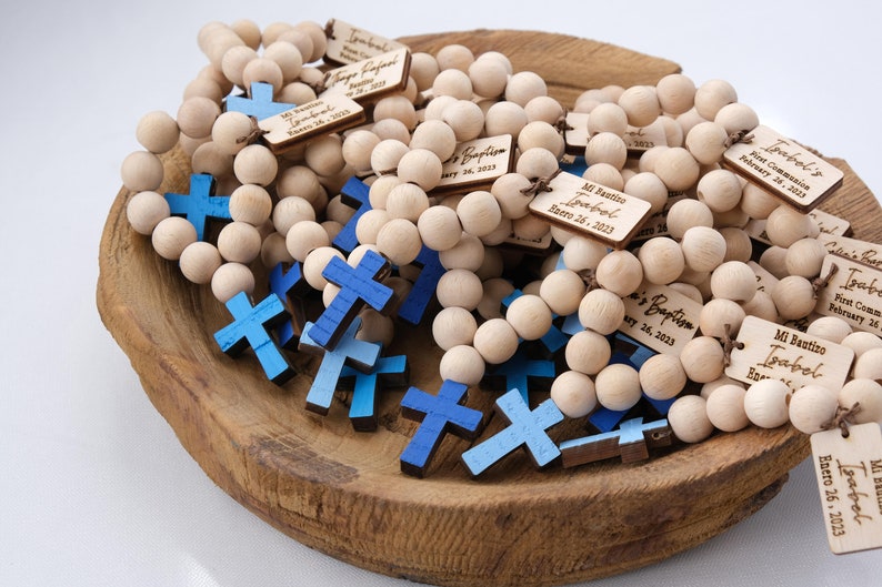 May include: Wooden bead rosaries with blue crosses and personalized tags. The tags are engraved with names and dates, likely for a baptism or first communion. The rosaries are displayed in a wooden bowl.