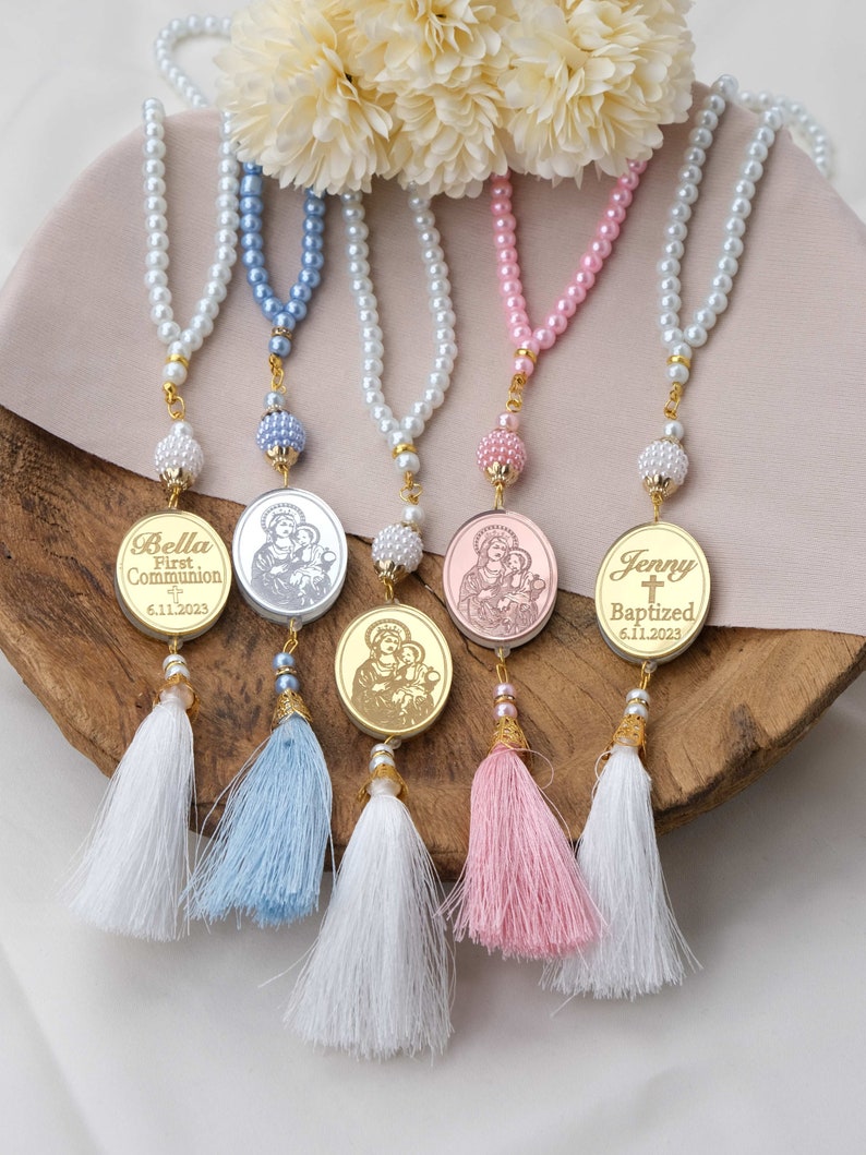 May include: Four pearl necklaces with gold tassels and personalized medallions. The medallions feature religious imagery and the names Bella, Jenny, and the dates 6.11.2023. The necklaces are in blue, white, pink, and silver.