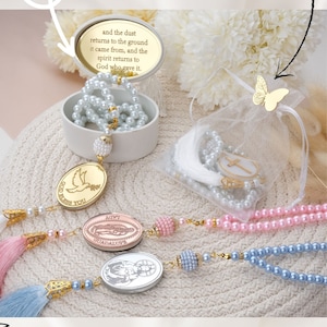 May include: A set of pearl prayer beads with a variety of charms, including a dove, a religious figure, and a butterfly. The beads are available in pink, blue, and white. The beads are packaged in a white box with a gold lid and a white organza bag with a gold butterfly charm. The text "God Bless You" is on the dove charm. The text "and the dust returns to the ground it came from, and the spirit returns to God who gave it." is on the lid of the box. The text "ALL PERSONALIZED COLORS TEXTS FIGURES" is at the bottom of the image.