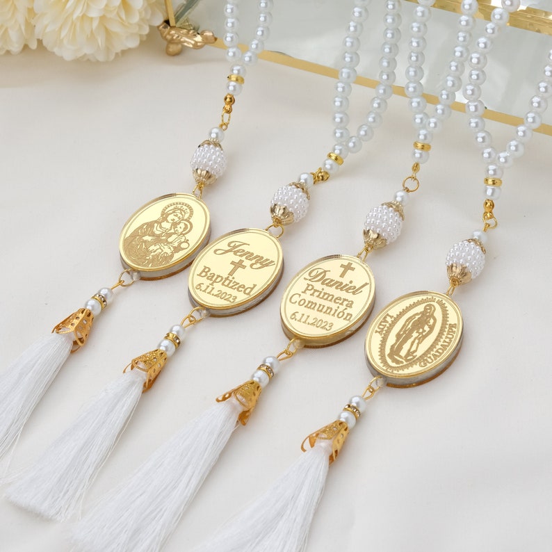 May include: Three gold oval-shaped charms with white tassels hanging from a white pearl necklace. The charms have a cross and text that reads "Jenny Baptized 6.11.2023", "Daniel Primera Comuni&oacute;n 6.11.2023", and "Nuestra Se&ntilde;ora de Guadalupe".