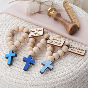 May include: Wooden rosary beads with light blue crosses and personalized tags. The tags read "Tiago Rafael Bautizo Enero 26, 2023", "Isabel's First Communion February 26, 2023", and "Sofia's Baptism February 26, 2023". A wooden stamp and a small gift box are also visible.