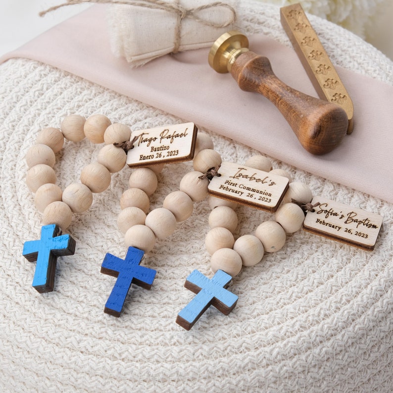 May include: Wooden rosary beads with light blue crosses and personalized tags. The tags read "Tiago Rafael Bautizo Enero 26, 2023", "Isabel's First Communion February 26, 2023", and "Sofia's Baptism February 26, 2023". A wooden stamp and a small gift box are also visible.