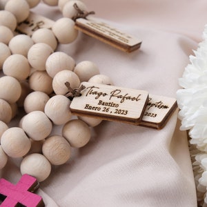 May include: Wooden bead rosary with personalized tags. The tags are engraved with the name "Tiago Rafael", the word "Bautizo", and the date "Enero 26, 2023". The rosary also features small wooden crosses in shades of purple and pink.