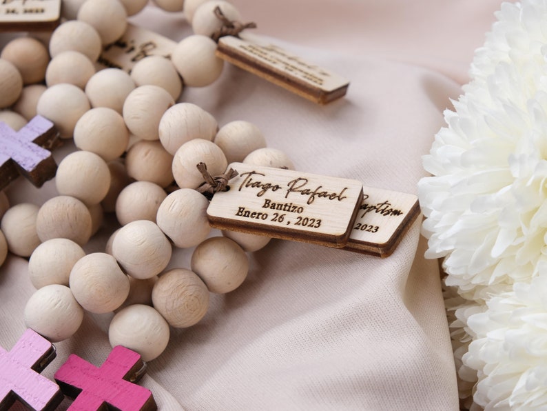 May include: Wooden bead rosary with personalized tags. The tags are engraved with the name "Tiago Rafael", the word "Bautizo", and the date "Enero 26, 2023". The rosary also features small wooden crosses in shades of purple and pink.