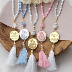 Puede incluir: Cuatro collares de perlas con borlas doradas y medallones personalizados. Los medallones presentan im&aacute;genes religiosas y los nombres Bella, Jenny, y las fechas 6.11.2023. Los collares son de color azul, blanco, rosa y plateado.
