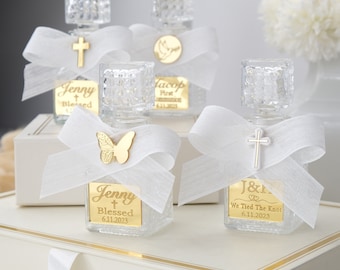 Puede incluir: Cuatro pequeñas botellas de vidrio con etiquetas doradas y lazos de cinta blanca. Las etiquetas tienen texto personalizado, incluidos nombres, fechas y frases como "Blessed" y "We Tied the Knot".