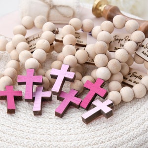 May include: Wooden rosary beads with small tags and pink crosses. The tags are engraved with names and dates, such as "Sofia's Baptism February 26, 2023." The crosses are in shades of pink. The beads are made of light-colored wood.