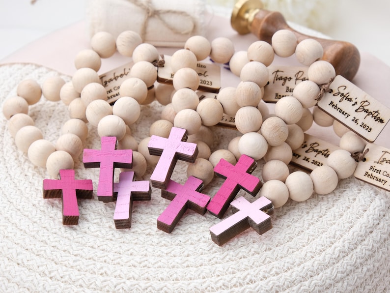 May include: Wooden rosary beads with small tags and pink crosses. The tags are engraved with names and dates, such as "Sofia's Baptism February 26, 2023." The crosses are in shades of pink. The beads are made of light-colored wood.