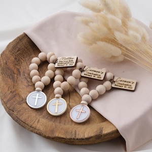 May include: Three wooden bead bracelets with a cross charm. Each bracelet has a personalised wooden tag with a name and the text "May your faith always guide you" and a date. The charms are silver, gold, and rose gold.