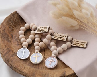 May include: Three wooden bead bracelets with a cross charm. Each bracelet has a personalized wooden tag with a name and the text "May your faith always guide you" and a date. The charms are silver, gold, and rose gold.