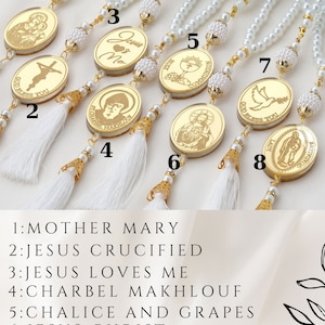 May include: Eight gold-colored oval charms with different religious figures and text. The charms are attached to white pearl necklaces with white tassels. The charms are numbered 1 through 8. The text below the charms reads: 1: MOTHER MARY, 2: JESUS CRUCIFIED, 3: JESUS LOVES ME, 4: CHARBEL MAKHLOUF, 5: CHALICE AND GRAPES, 6: JESUS CHRIST, 7: DOVE, 8: OUR LADY OF GUADALUPE.