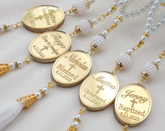 May include: Five gold oval-shaped charms with white tassels and pearl beads. Each charm has a name, a cross, and a date engraved on it. The names are Matteo, Fiago, Nicholas, Matteo, and Jenny. The dates are 6.11.2023. The charms are for a baptism or christening.