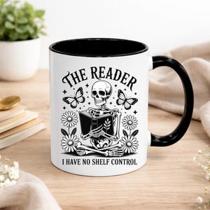 Puede incluir: Taza de cerámica blanca con interior y asa negros. La taza presenta una ilustración en blanco y negro de un esqueleto leyendo un libro, con el texto "The Reader" y "I Have No Shelf Control".