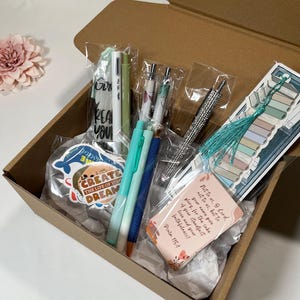 May include: A brown cardboard gift box containing several items: a bookmark with a teal tassel, a rhinestone-encrusted pen, two blue and green pens, a white pen, a sticker reading "CREATE THE LIFE OF YOUR DREAMS," and a card with Psalm 115:1 text.