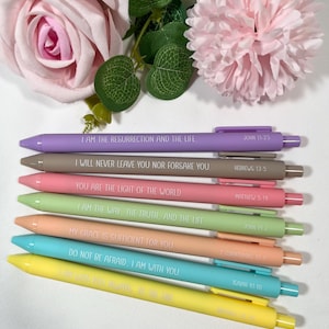 May include: A collection of seven colorful pens with inspirational quotes printed on them. The pens are in shades of purple, brown, pink, green, peach, blue, and yellow. Each pen has a different quote and a corresponding Bible verse reference.