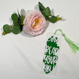 May include: A clear acrylic bookmark with the text "Girl, Read Your Bible" in white over a green shape. It has a green tassel and is next to a pink rose and green leaves.