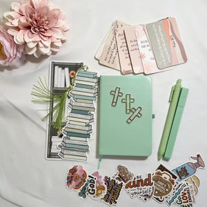 May include: A light green journal with three cross icons, a bookmark featuring a stack of books, two light green pens, several scripture cards, and various faith-themed stickers. The set includes pink artificial flowers.