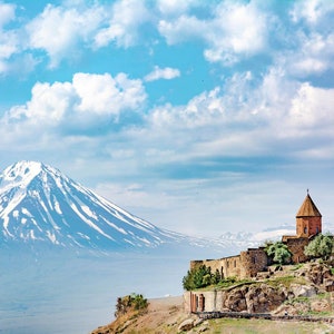 May include: A scenic landscape featuring a snow-capped mountain under a cloudy blue sky. A historic stone structure with a conical roof sits on a cliffside, overlooking the valley. The image captures a serene, natural environment.