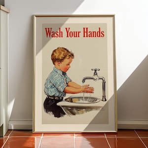 Puede incluir: Una lámina artística enmarcada con una ilustración de estilo vintage de un niño lavándose las manos en un lavabo. El texto "Wash Your Hands" aparece impreso en rojo en la parte superior. La obra utiliza una paleta de colores suaves con fondo crema.