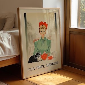 May include: A framed poster featuring an illustration of a person with blonde hair wearing a red headpiece and a green blouse. A black and white cat sits beside a red teapot and teacup. The text reads, "TEA FIRST, DARLING" at the bottom.